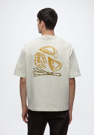 Homme portant un t-shirt beige clair avec des dessins linéaires jaunes de fruits de mer et d'oignons verts dans le dos, debout devant un fond uni.