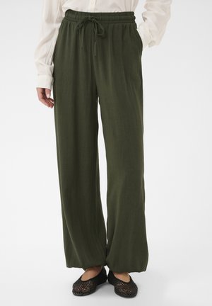 Kobieta w luźnych oliwkowozielonych spodniach z sznurkiem w pasie, białej bluzce z długim rękawem i czarnych plecionych wsuwanych butach.