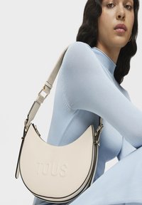 Bolso de hombro en forma de media luna de cuero blanco con logo "TOUS" en relieve, ribete negro y correa desmontable. El modelo lleva una blusa de color azul claro.