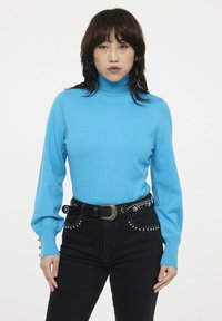 Pull bleu à col roulé en maille lisse avec des manches longues. Associé à un jean noir avec des accents cloutés et une ceinture épaisse.