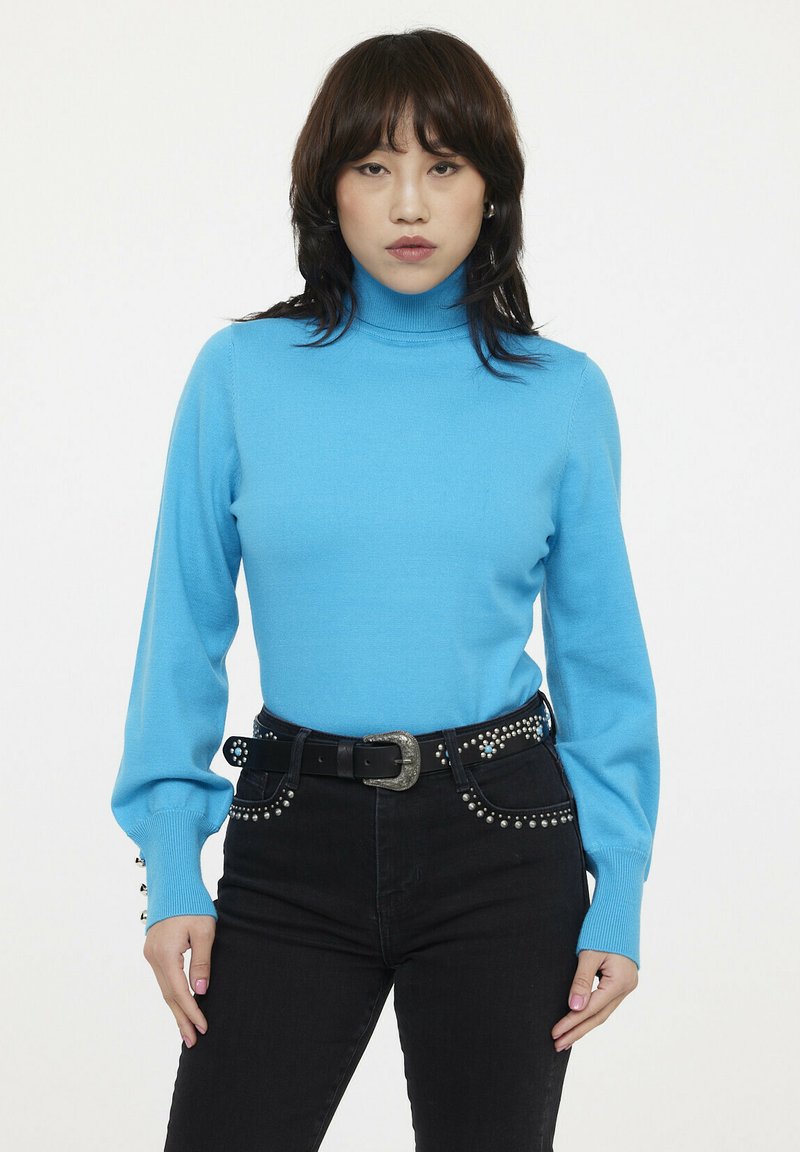 Pull bleu à col roulé en maille lisse avec des manches longues. Associé à un jean noir avec des accents cloutés et une ceinture épaisse.