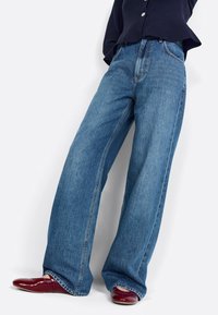 Wide-leg jeans in medium blue denim, featuring a high waist, front seams, and subtle whiskering. Paired with red pointed shoes.