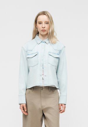 Femme blonde de face portant une chemise en denim bleu clair à boutons avec poches poitrine et un pantalon beige taille haute, fond neutre.