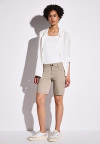 Shorts beiges au design ajusté et poches avant, associés à un débardeur blanc et une chemise blanche ouverte. Des baskets blanches complètent le look.