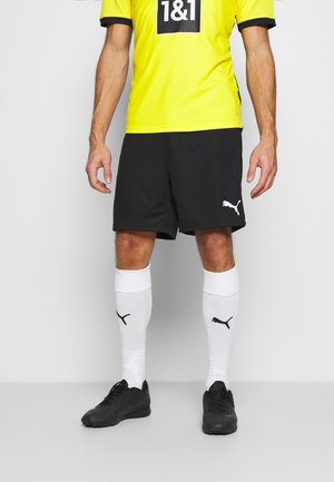 Gelbes Sporttrikot mit schwarzen Akzenten, kombiniert mit schwarzen Shorts. Weiße Socken mit schwarzem Logo und schwarze Fußballschuhe. Einfaches sportliches Design.