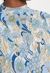 Långärmad topp med hög krage, tillverkad av textil med struktur. Blommönster i nyanser av blått, vitt och guldaccenter.