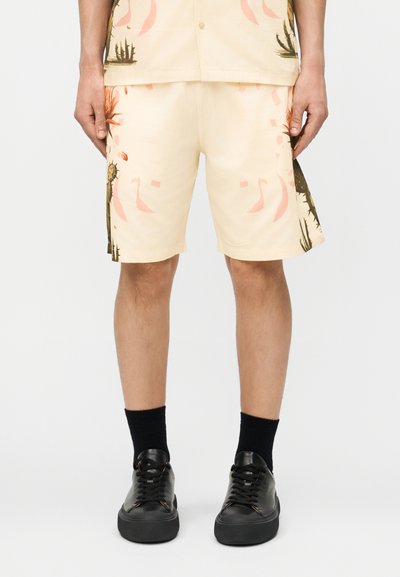 Ανδρικό μπεζ σορτς με αφηρημένα ροζ και σκούρα πράσινα βοτανικά μοτίβα, συνδυασμένο με μαύρες κάλτσες και μαύρα δερμάτινα παπούτσια με κορδόνια.