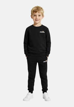 Jeune garçon debout, portant un sweat-shirt Ellessee noir et un pantalon de survêtement assorti avec des baskets blanches, les mains dans les poches, souriant.