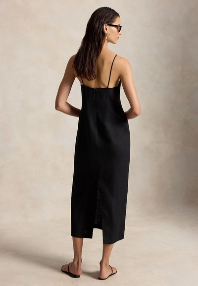 Γυναίκα με μακριά σκούρα μαλλιά που φοράει μαύρο μίντι φόρεμα χωρίς μανίκια με λεπτές τιράντες, μαύρα σανδάλια και μαύρα γυαλιά, στέκεται μπροστά σε απλό φόντο.