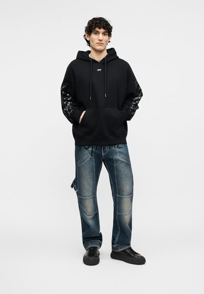Jauns vīrietis, valkājot melnu kapuci ar grafiku uz piedurknēm un nodilušas zilas džinsas, stāv ar rokām kabatās pret vienkāršu fonu.
