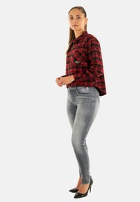 Giovane donna in giacca a quadri rossi e neri, jeans skinny grigi e tacchi alti neri, in piedi di lato su sfondo bianco.