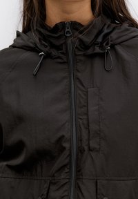 Giacca nera leggera con chiusura a zip, cappuccio regolabile e tasca sul petto. Tessuto liscio con un design semplice e minimalista.
