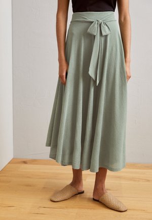 Mujer que lleva una falda midi verde claro con un lazo en la cintura, un top negro y zapatos beige tejidos sin cordones, de pie sobre un suelo de madera.