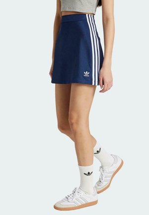 Falda de tenis azul con franjas laterales blancas, con una cinturilla elástica. La modelo lleva zapatillas blancas y calcetines con acentos de logo negro.