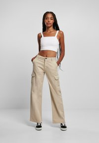 Pantalones cargo beige de pierna ancha con bolsillos laterales, combinados con una camiseta de tirantes blanca corta y zapatillas negras, con cierre de botón y corte relajado.