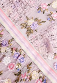 Embroidered lace fabric in lavender with floral patterns. Features multicolored thread accents and a pastel striped trim. Soft texture.