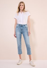 Jean bleu clair, taille mi-haute, avec un design de jambe droite raccourci et une texture décolorée, associé à une chemise blanche à boutons et des baskets blanches.