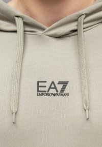 Mikina s kapucňou v svetlo béžovej látke s hladkou textúrou. Má čierne logo EA7 Emporio Armani a opletené šnúrky na sťahovanie.