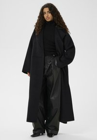 Cămașă oversized neagră din lână cu rever larg, asortată cu un pulover negru cu guler înalt și pantaloni largi din piele neagră lucioasă. Pantofi negri cu vârful ascuțit.