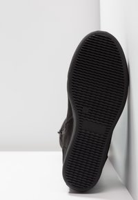 Chaussure en daim noir avec une semelle en caoutchouc texturée, présentant un design plat et une fermeture éclair latérale. La semelle extérieure comporte un motif ondulé pour une meilleure adhérence.