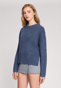 Niebieski sweter z dzianiny z ribbowanym wykończeniem, okrągłym dekoltem i bocznymi rozcięciami. Noszony na szarych szortach, charakteryzujących się miękką teksturą i luźnym fasonem.