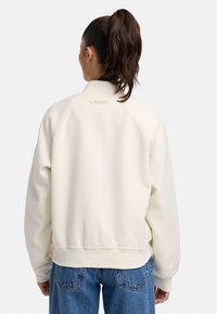 Crème-kleurige sweatshirt met ribgebreide kraag en manchetten, voorzien van een subtiel merkembleem op de achterkant. Gecombineerd met blauwe jeans.