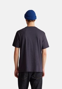 Osoba v tmavě šedém tričku s krátkým rukávem, černých džínách a modré pletené čepici, otočená zády na jednobarevném bílém pozadí.