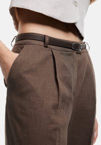 Bruine linnen broek met plooien en een slanke bruine leren riem met een zilveren gesp, tentoongesteld op taillehoogte met een hand in de zak.