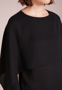 Indumento nero con scollatura rotonda e maniche a mantello fluenti. Tessuto liscio con un leggero lucido, caratterizzato da elementi di design a strati.