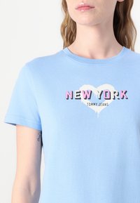 Šviesiai mėlyna medvilninė marškinėliai trumpomis rankovėmis, su spausdinta grafika, kurioje rašoma "NEW YORK" juoda ir rožine spalvomis širdies dizaino fone.