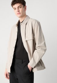 Light beige jacket with a collar, button closure, and chest pocket, paired with a black polo shirt and black trousers. Smooth fabric texture.