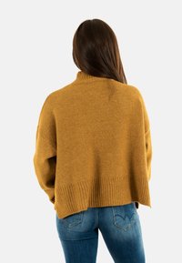 Un pull surdimensionné jaune moutarde avec un col côtelé haut et un ourlet côtelé, fabriqué à partir d'un matériau doux. Porté avec un jean bleu ajusté.