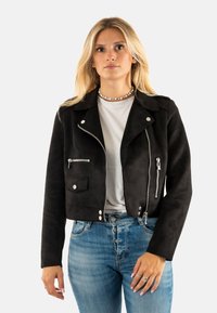 Chaqueta corta de ante negro con cierre frontal, herrajes plateados, bolsillos laterales y un diseño clásico de cuello. Combinada con jeans azules.