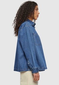 Blauwe spijkerblouse met een ontspannen pasvorm, voorzien van een kraag, voorzak en knoopmanchetten. De stof heeft een klassieke spijkertextuur.
