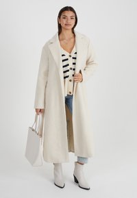 Cappotto lungo color crema con finitura testurizzata, colletto a punte e tasche laterali. Abbinato a un cardigan a righe, jeans chiari e stivali alla caviglia beige.