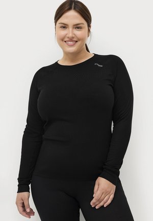 Kvinde med brunt hår samlet bagud, smilende, iført en tætsiddende sort langærmet trøje og sorte bukser foran en ensartet hvid baggrund.