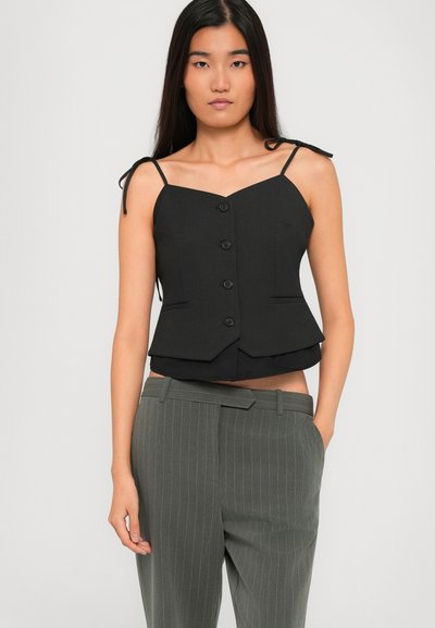 Woman wearing black buttoned sleeveless top with tie straps and gray pinstriped pants, standing against plain light background.