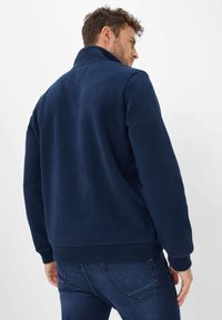 Giacca in fleece blu navy con colletto alto, polsini a costine e una texture unita. Progettata per una vestibilità comoda, presenta dettagli di cucitura minimi.