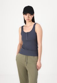 Grå, ribbestrikket tanktop med utringet hals, med knapper. Kombinert med avslappede olivenfargede joggebukser og en svart caps.
