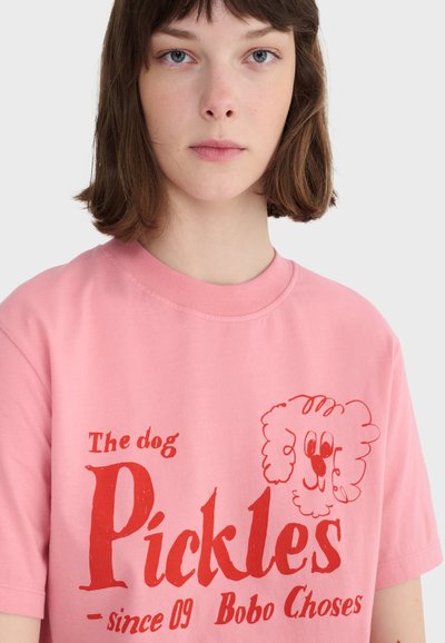 Mladý človek s krátkymi hnedými vlasmi, oblečený v ružovom tričku s červeným nápisom a kresleným psíkom na prednej strane.
