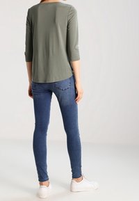 Grünes Dreiviertelarm-Oberteil, kombiniert mit blauen Skinny-Jeans und weißen Sneakers; besteht aus glattem Stoff und hat ein einfaches, klassisches Design.