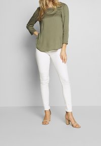 Top khaki a maniche lunghe con scollatura rotonda, abbinato a jeans skinny bianchi e sandali con tacco a blocco beige. Tessuto liscio con una leggera lucentezza.