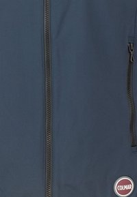 Giacca softshell blu navy con chiusura frontale a cerniera, tessuto testurizzato e tasca laterale. Presenta un logo rotondo in un colore a contrasto.