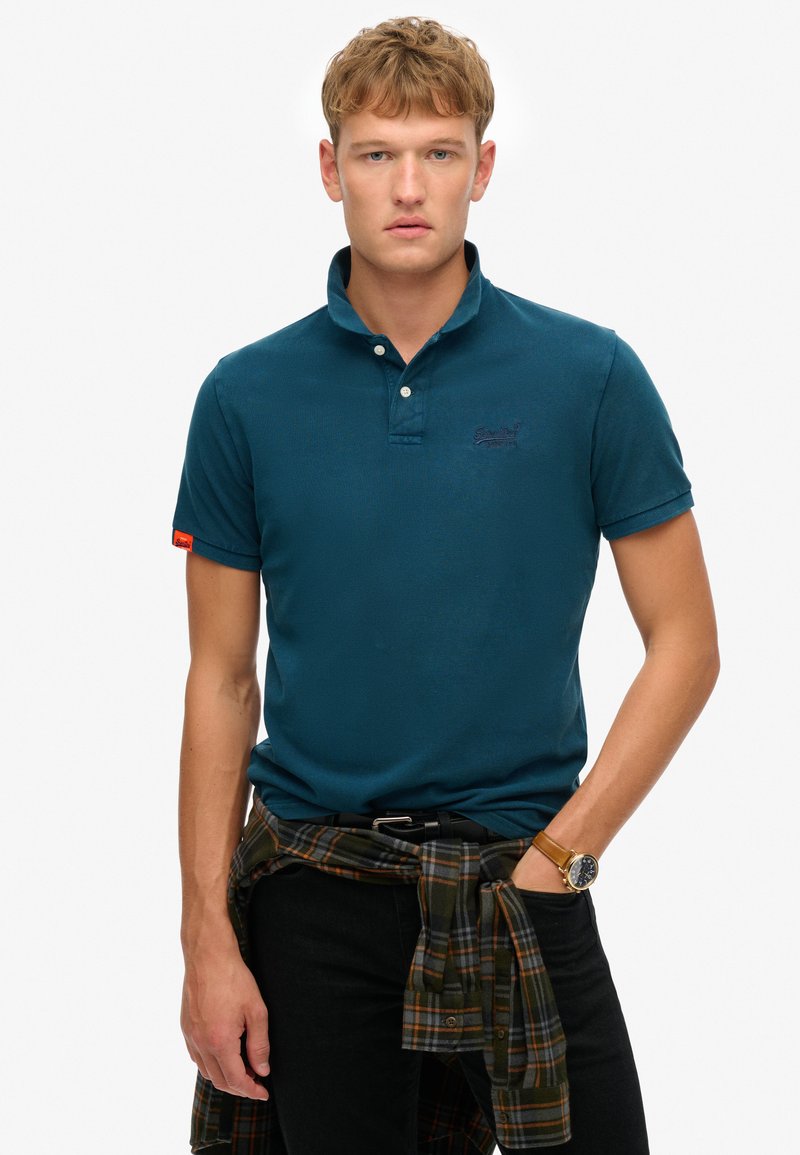 Polo shirt color teal con un tessuto texturizzato, colletto a due bottoni, logo ricamato e maniche corte. Jeans neri con una camicia a quadri annodata in vita.