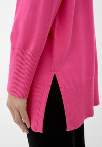 Rosa gestrickter Pullover mit gerippten Ärmeln und Saum. Verfügt über einen seitlichen Schlitz am unteren Rand, der eine glatte Textur und eine lockere Passform zeigt.