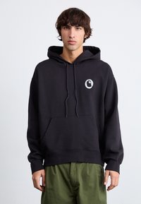 Sudadera negra con bolsillo frontal, cordones y un logo redondo en el pecho. Material suave de algodón con un corte holgado.