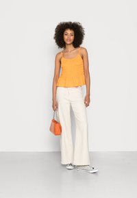 Top peplum em laranja com pregas, com alças finas, combinado com calças cream de boca de sino. Acessórios incluem uma pequena mala laranja e ténis preto e branco.