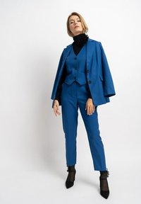 Blauer Blazer mit Cape-Detail, passende taillierte Hose und schwarzer Rollkragenpullover. Schwarze High Heels mit transparenten Socken. Glatter Stoff, maßgeschneiderter Schnitt.