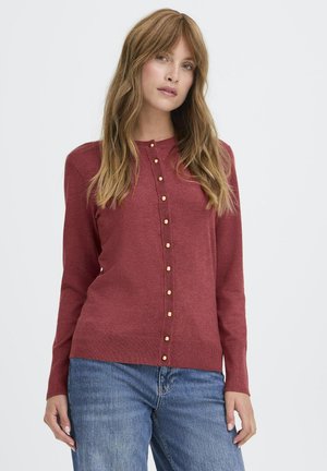 Rote Strickjacke mit langen Ärmeln, rundem Ausschnitt und Knopfleiste mit goldenen Knöpfen. Kombiniert mit blauen Jeans.