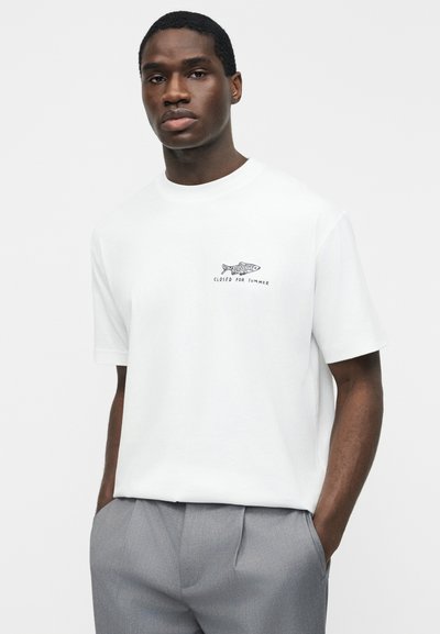 Jeune homme portant un t-shirt blanc avec un petit motif de poisson et le texte « Fermé pour l'été », associé à un pantalon gris, debout les mains dans les poches.